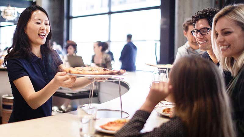 At a restaurant, a young woman serves a pizza to a smiling family.